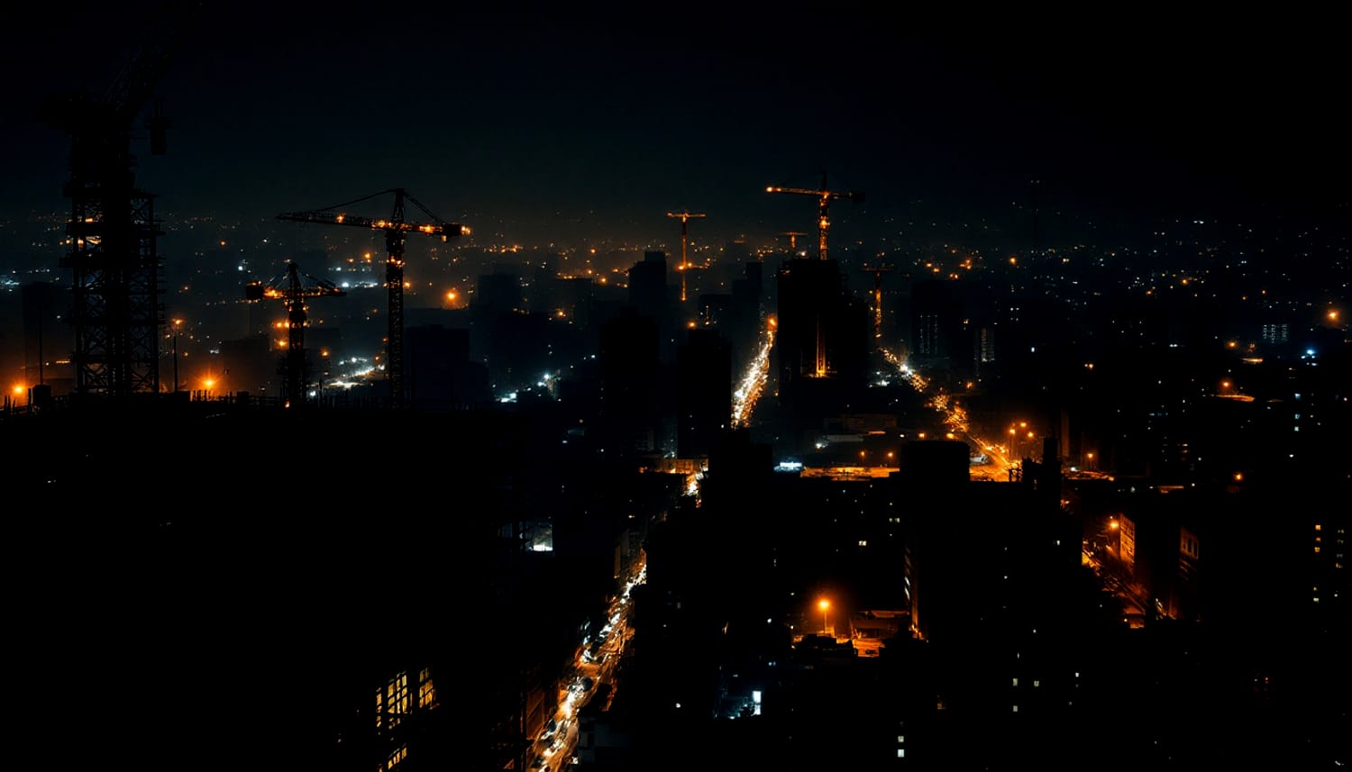 A city skyline at night with several tower cranes and clusters of work lights.